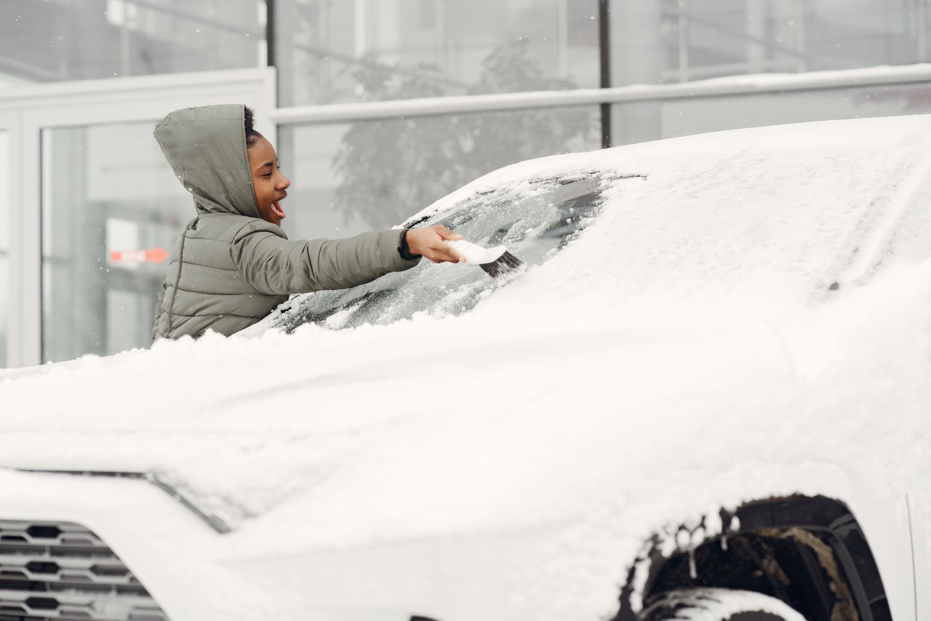 A woman in a gray coat clears snow from a car windshield on a cold winter day.